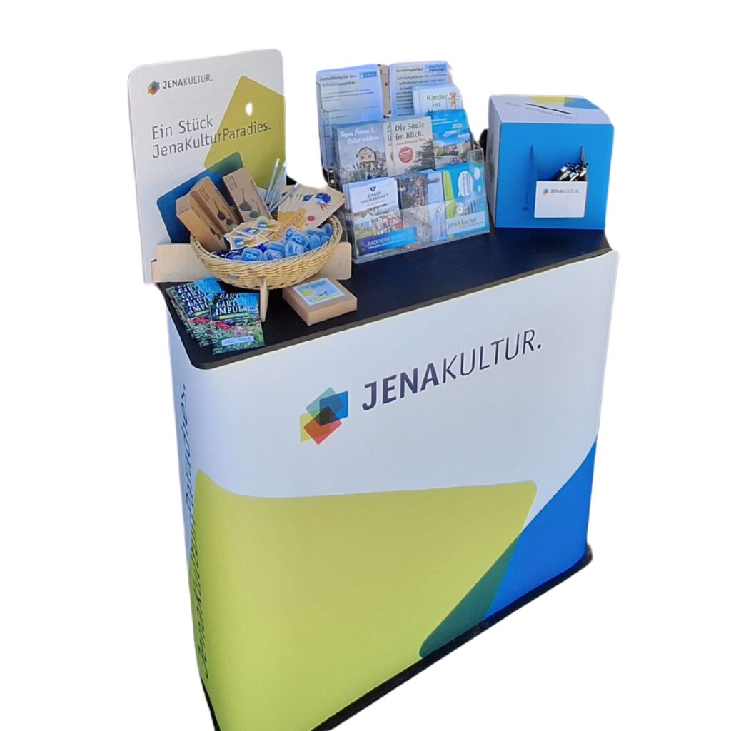Ein Tresen mit JenaKultur-Logo und -Design, auf dem ein Merchkorb, ein Flyerständer sowie eine Losbox stehen.
