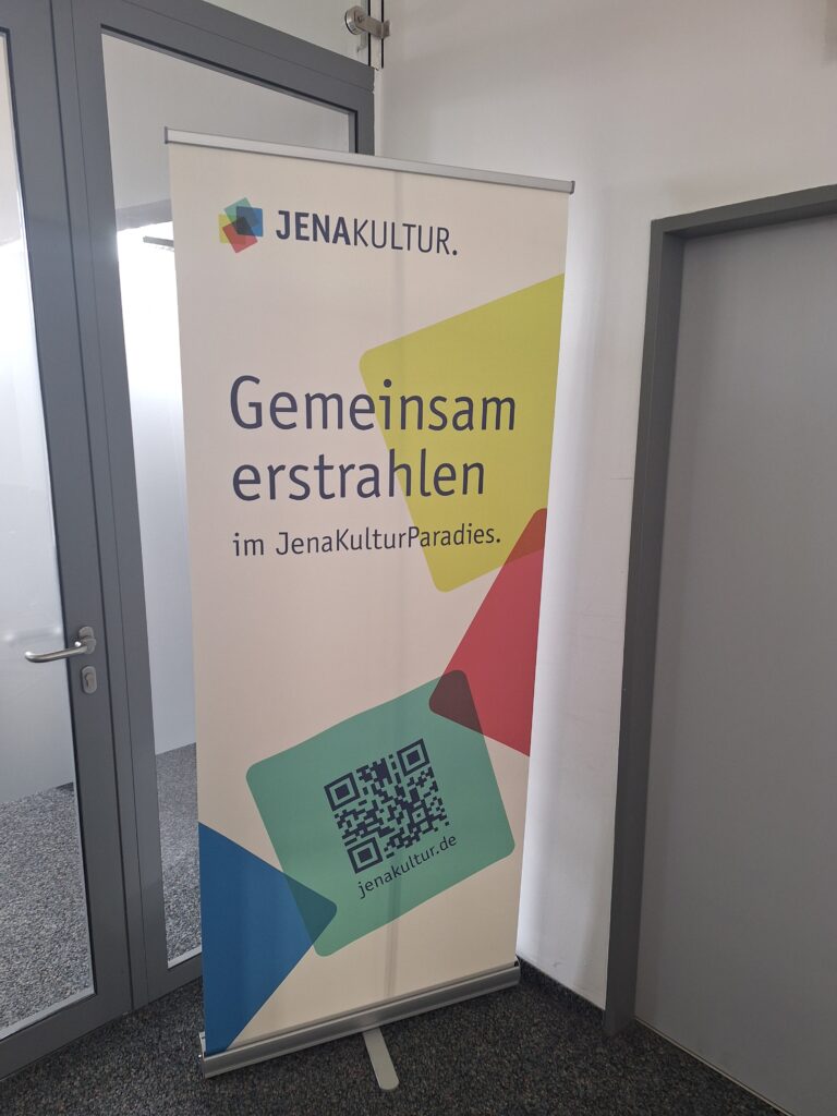 Rollup mit JenaKultur-Logo, das aus mehrfarbigen Quadraten besteht, mit dem Schriftzug "Gemeinsam erstrahlen im JenaKulturParadies" und einem QR-Code zur JenaKultur-Webseite.