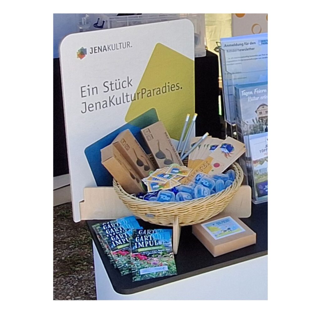 Ein Körbchen, in dem Merchandise liegt und auf dessen Display "Ein Stück JenaKulturParadies" steht und ein blauer und grüner Würfel sowie ein JenaKultur-Logo abgebildet sind.