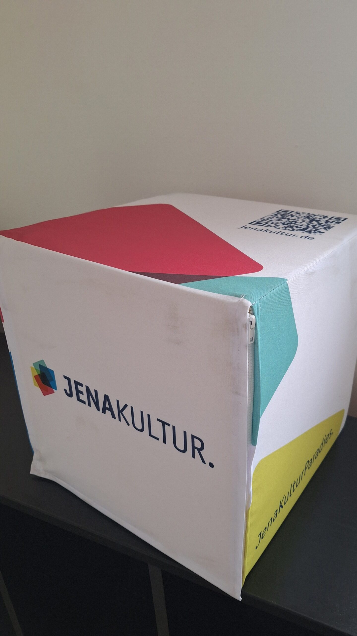 Ein mit weißem Stoff bezogener Sitzwürfel, auf dem farbige Quadrate, das JenaKultur-Logo und ein QR-Code abgebildet sind.